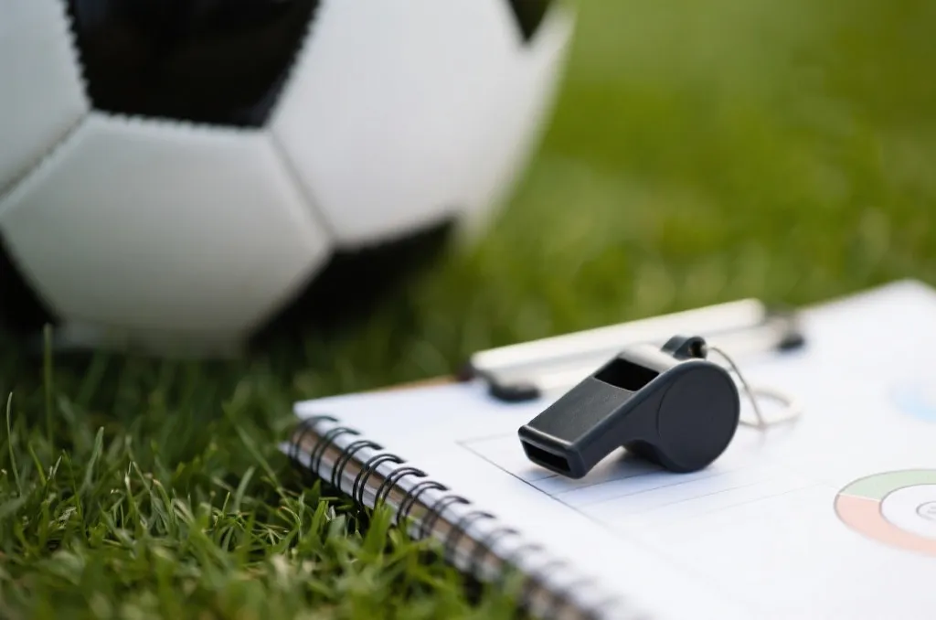 A close-up of a soccer referee's whistle resting on a tactical notebook next to a soccer ball on grass, shallow depth of field, editorial sports photography